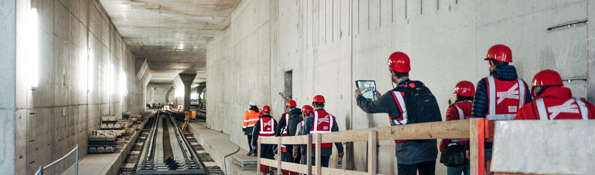 Menschen mit roten Helmen und Westen laufen durch einen großen, unfertigen U-Bahn-Tunnel.