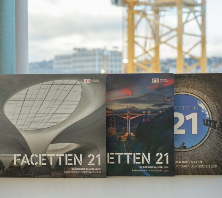 Drei "Facetten 21"-Bücher mit architektonischen Einbänden auf einer weißen Fläche neben einem Fenster mit Kränen draußen.