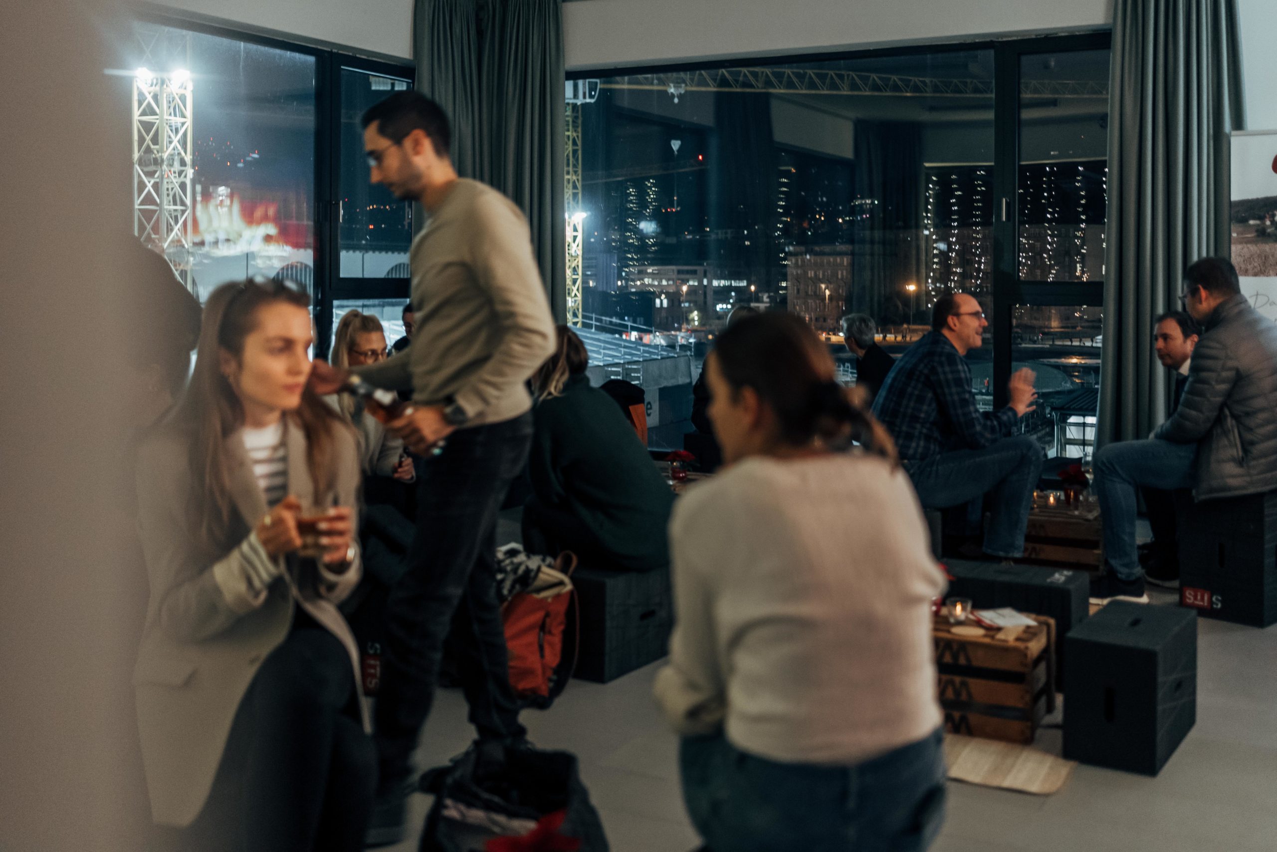 Menschen, die nachts in einem modernen Innenraum bei Getränken zusammenkommen, während draußen die Lichter der Stadt zu sehen sind.