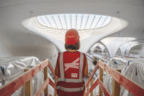 Person mit rotem Bauhelm und Warnweste auf Holzsteg bei STUTTGART 21, gebogene Architektur, Oberlicht.