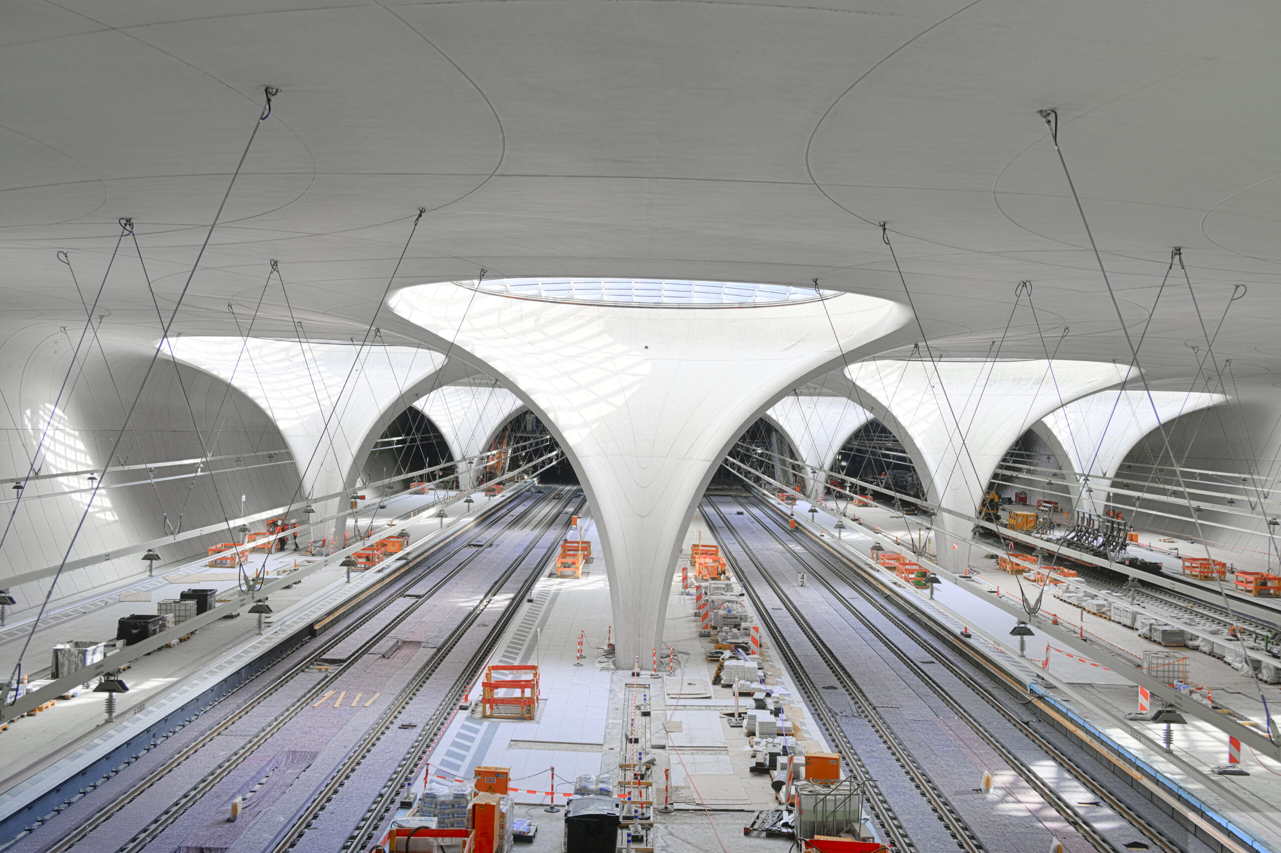 Moderner, im Bau befindlicher Bahnhof mit gebogenen weißen Decken, Gleisen und Bauabsperrungen.