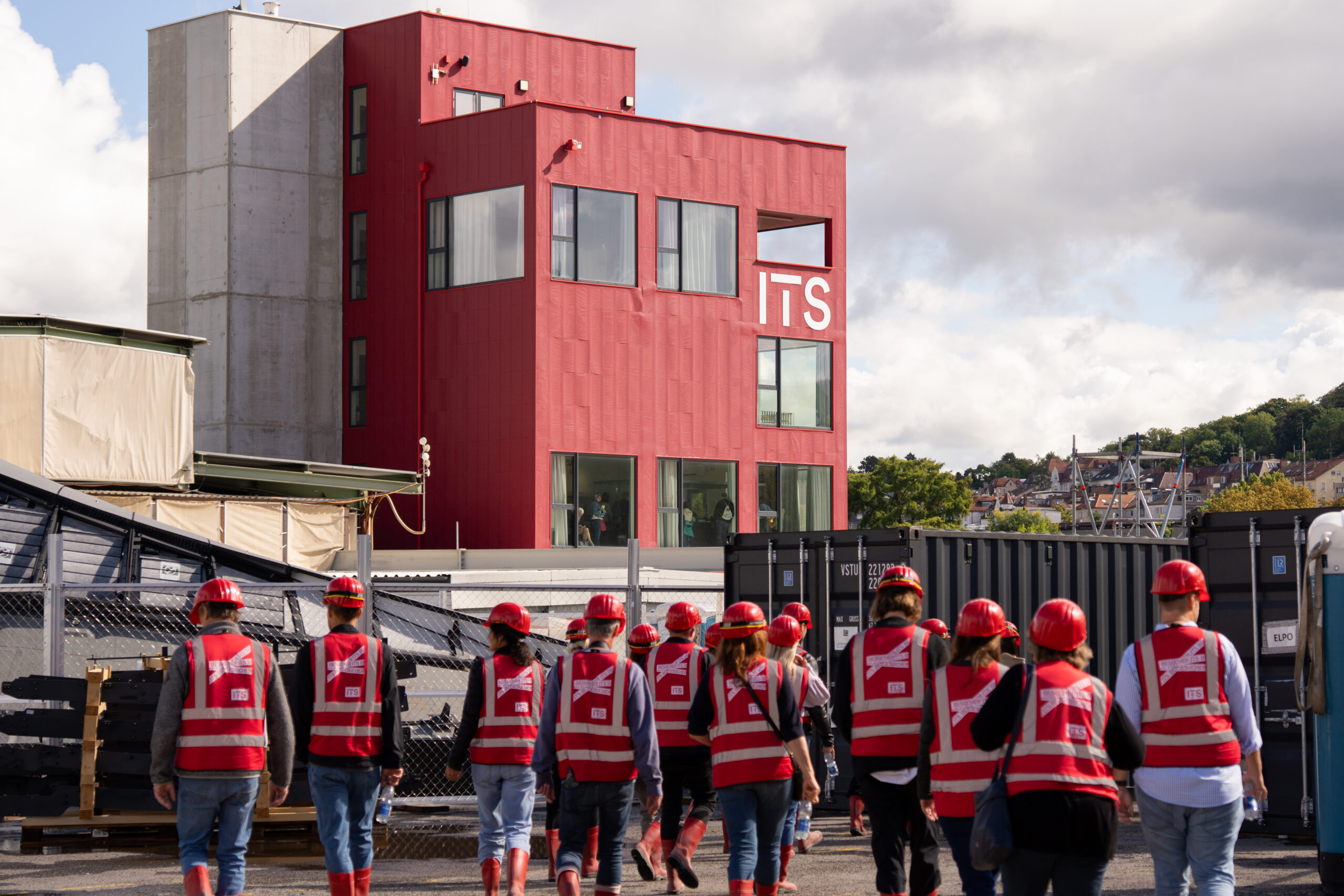 Menschen in roten Westen und Schutzhelmen gehen zu einem modernen roten "ITS"-Gebäude auf einer Baustelle.